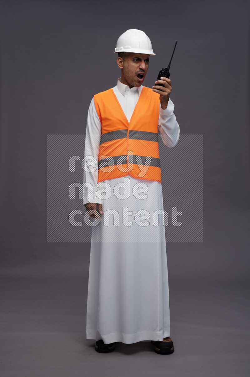Saudi man wearing thob with engineer vest standing holding walkie-talkie on gray background