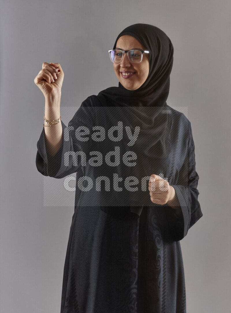 Woman in black abaya, hijab and glasses holding a marker pen to write on a transparent board with different reactions and expressions on grey background
