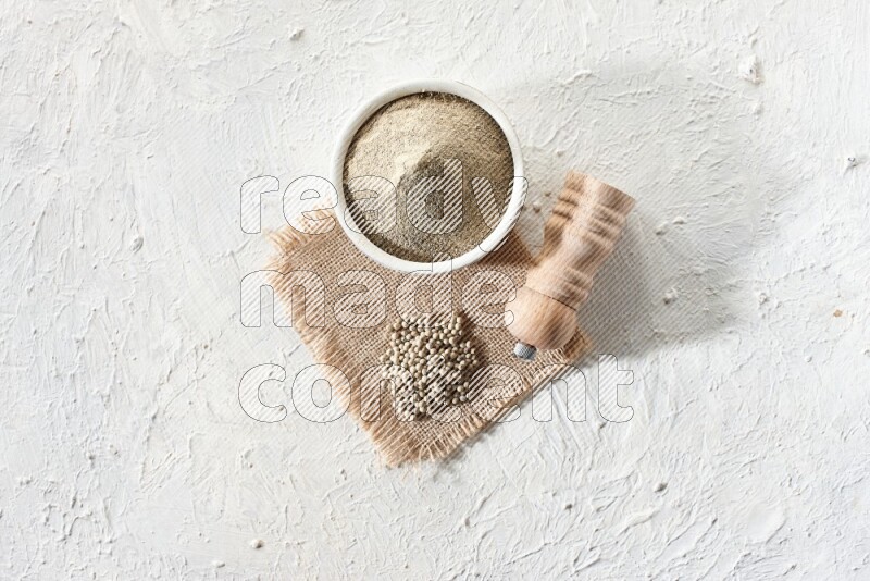 White pottery bowl full of white pepper powder set on a burlap piece of fabric with pepper beads and wooden pepper grinder on textured white flooring