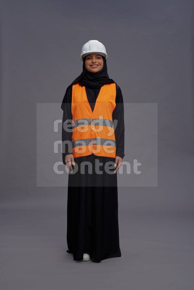 Saudi woman wearing Abaya with engineer vest standing interacting with the camera on gray background