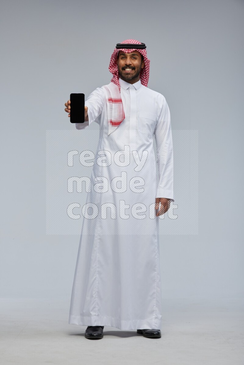 Saudi man Wearing Thob and shomag standing showing phone to camera on Gray background