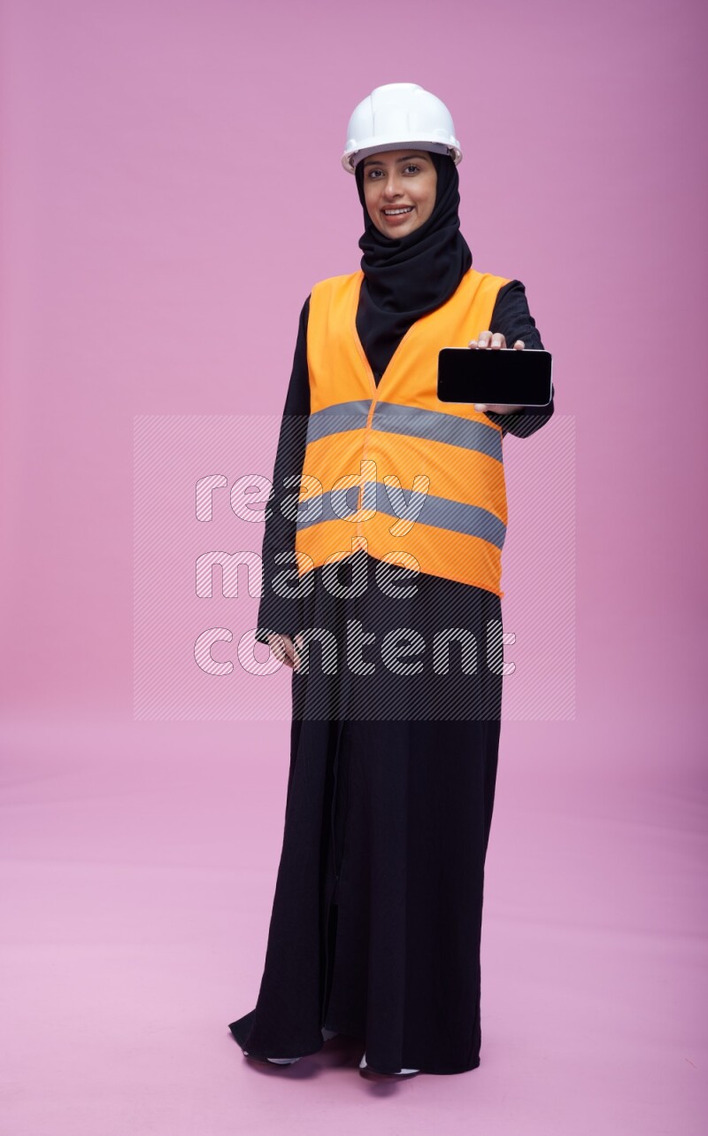 Saudi woman wearing Abaya with engineer vest and helmet standing showing phone to camera on pink background