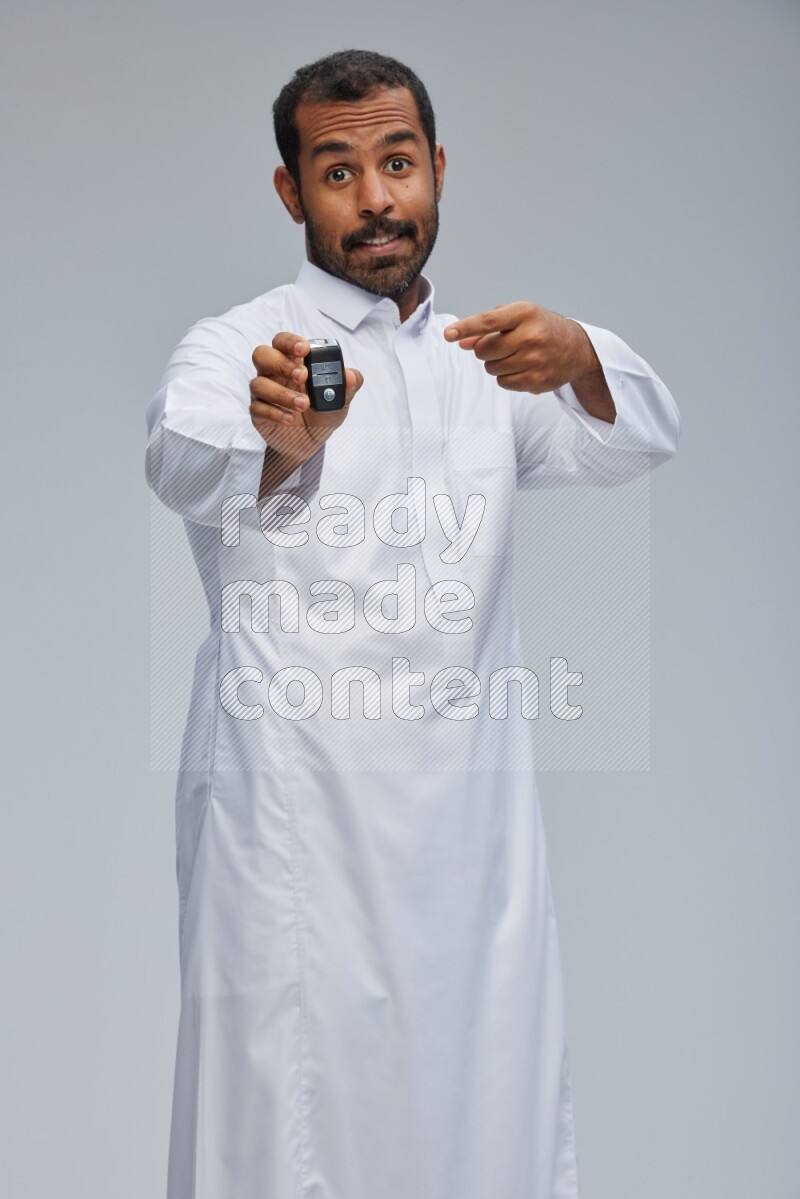 Saudi man wearing thob and shomag standing holding car key on gray background