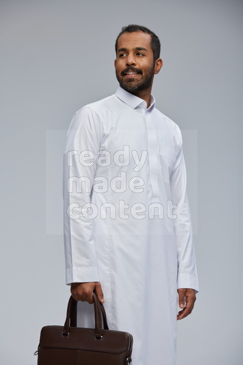 Saudi man wearing thob standing holding suitcase on gray background