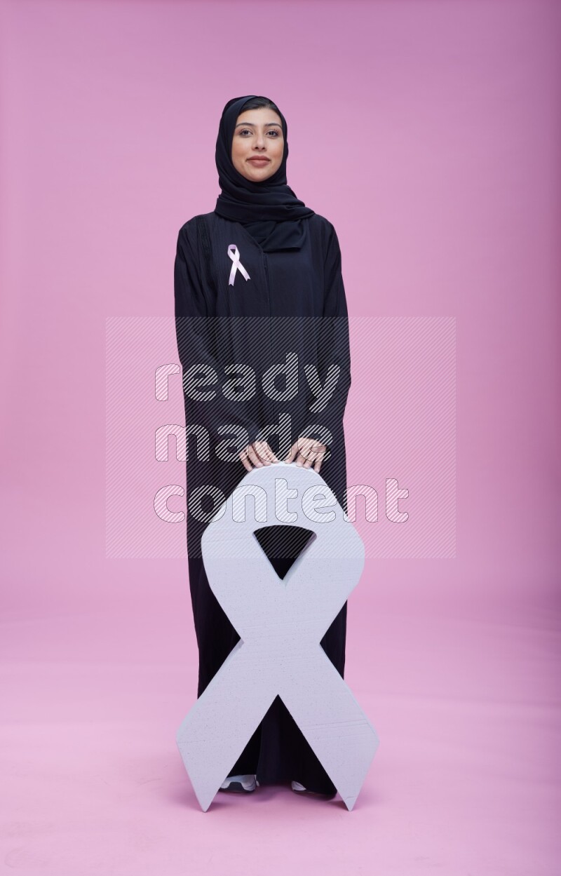 Saudi woman wearing pink ribbon on abaya standing holding awareness ribbon on plain pink background