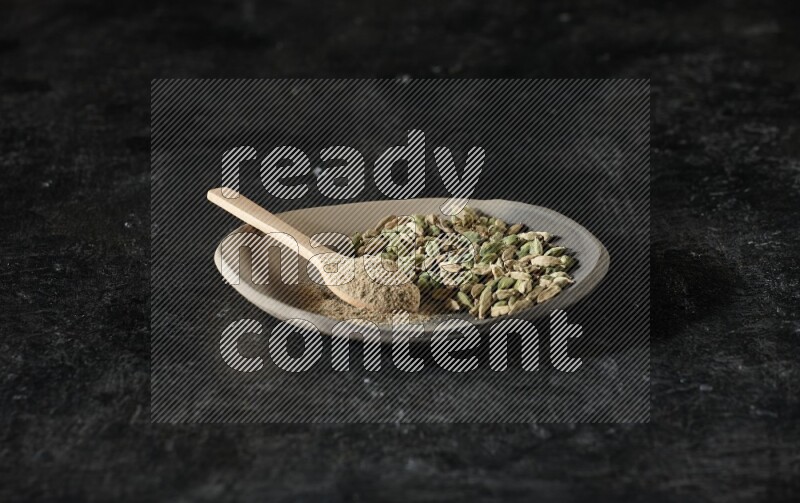 A plate filled with cardamom seeds and a wooden spoon full of cardamom powder on a textured black flooring