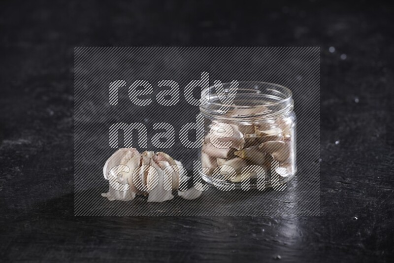 A glass jar full of garlic cloves with a whole garlic bulb beside it on a textured black flooring