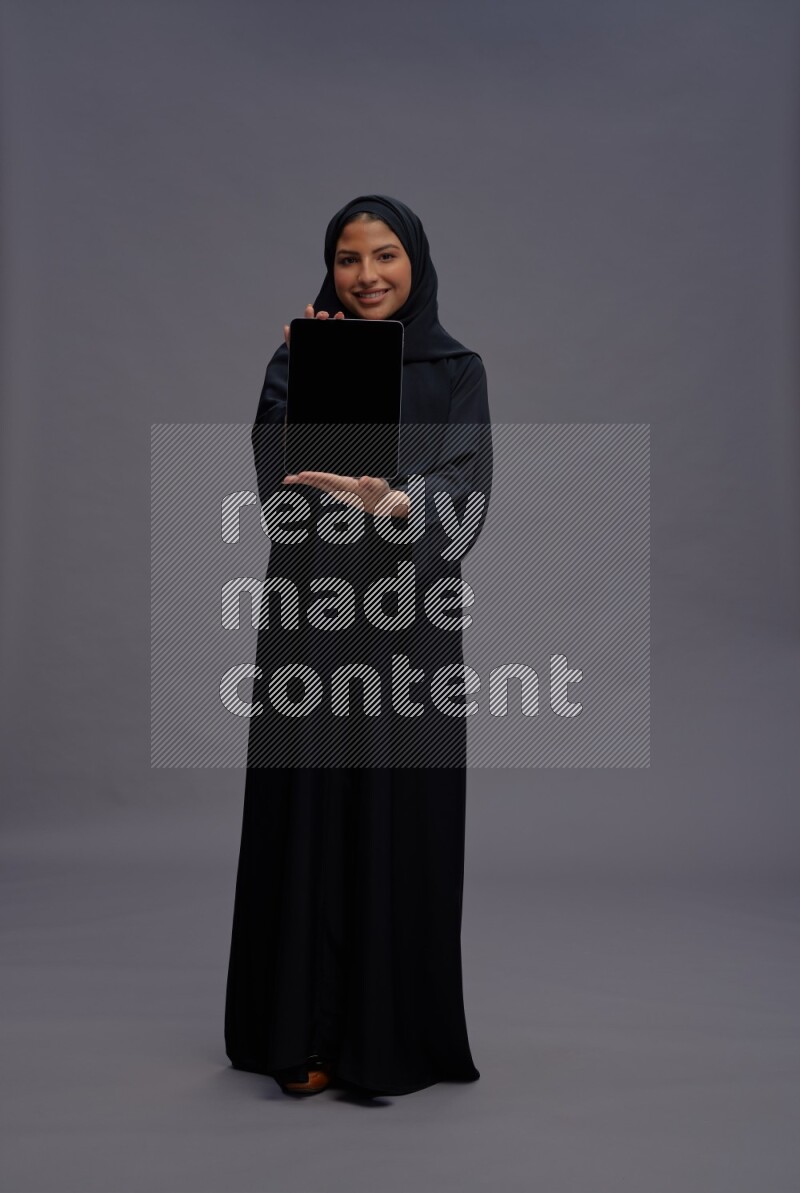 Saudi woman wearing Abaya standing showing tablet to camera on gray background