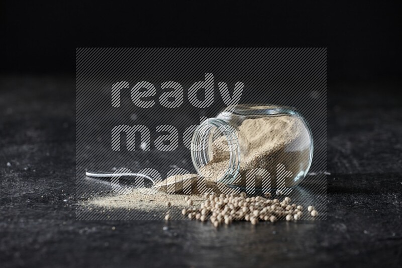 A flipped herbal glass jar and metal spoon full of white pepper powder with spilled powder and pepper beads on textured black flooring