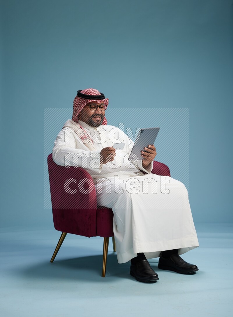 Saudi Man with shimag sitting on chair holding ATM card while working on tablet on blue background