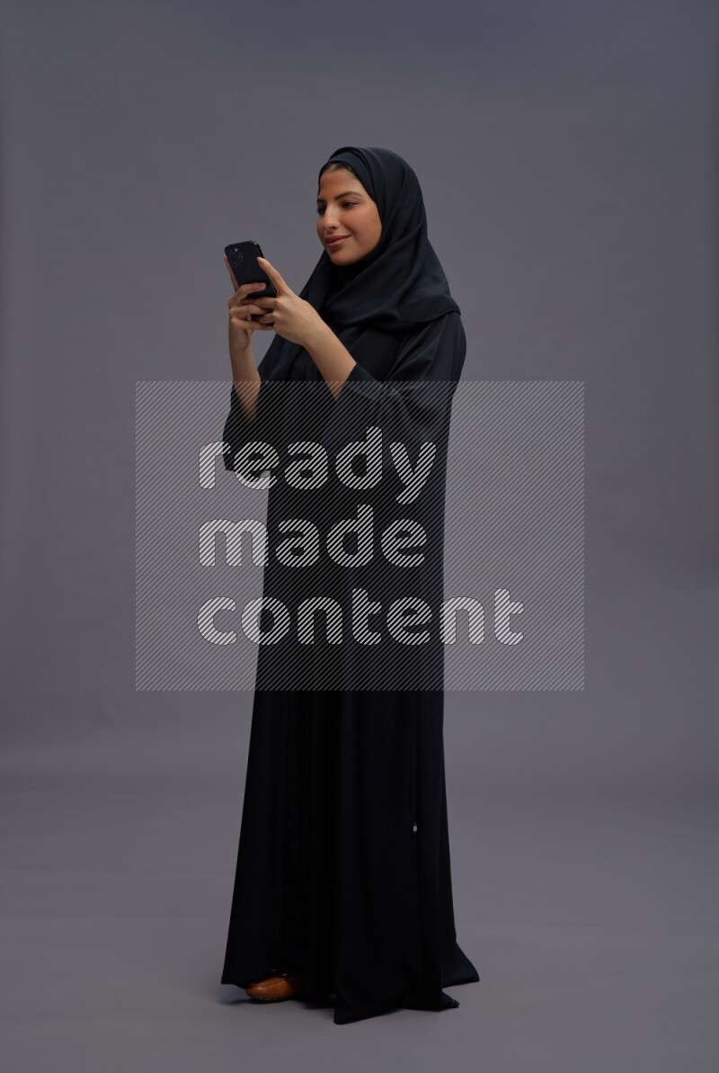 Saudi woman wearing Abaya standing texting on phone on gray background