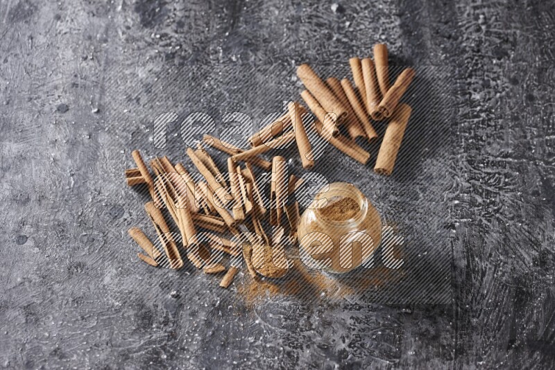 Herbal glass jar and a metal spoon full of cinnamon powder surrounded by cinnamon sticks on textured black background