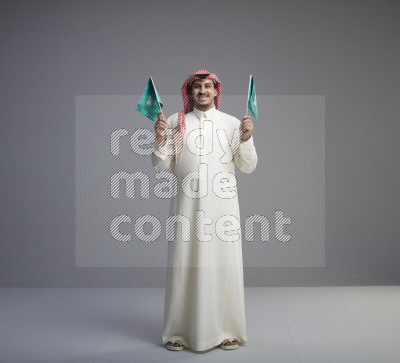 A Saudi man standing wearing thob and red shomag raising small Saudi flag on gray background