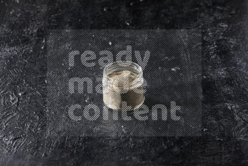 A glass jar full of white pepper powder on textured black flooring