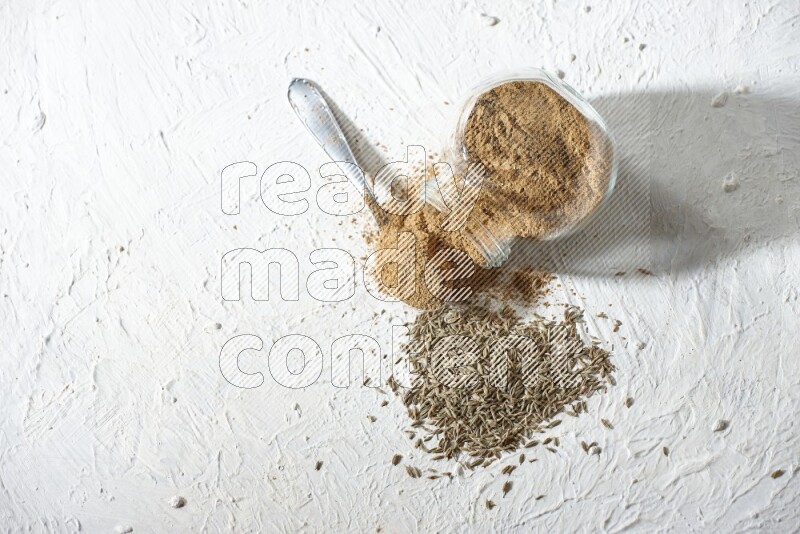 A glass spice jar and metal spoon full of cumin powder and the jar flipped and powder spilled out with cumin seeds on textured white flooring