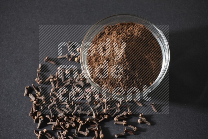 A glass bowl full of cloves powder with cloves grains spread on black flooring