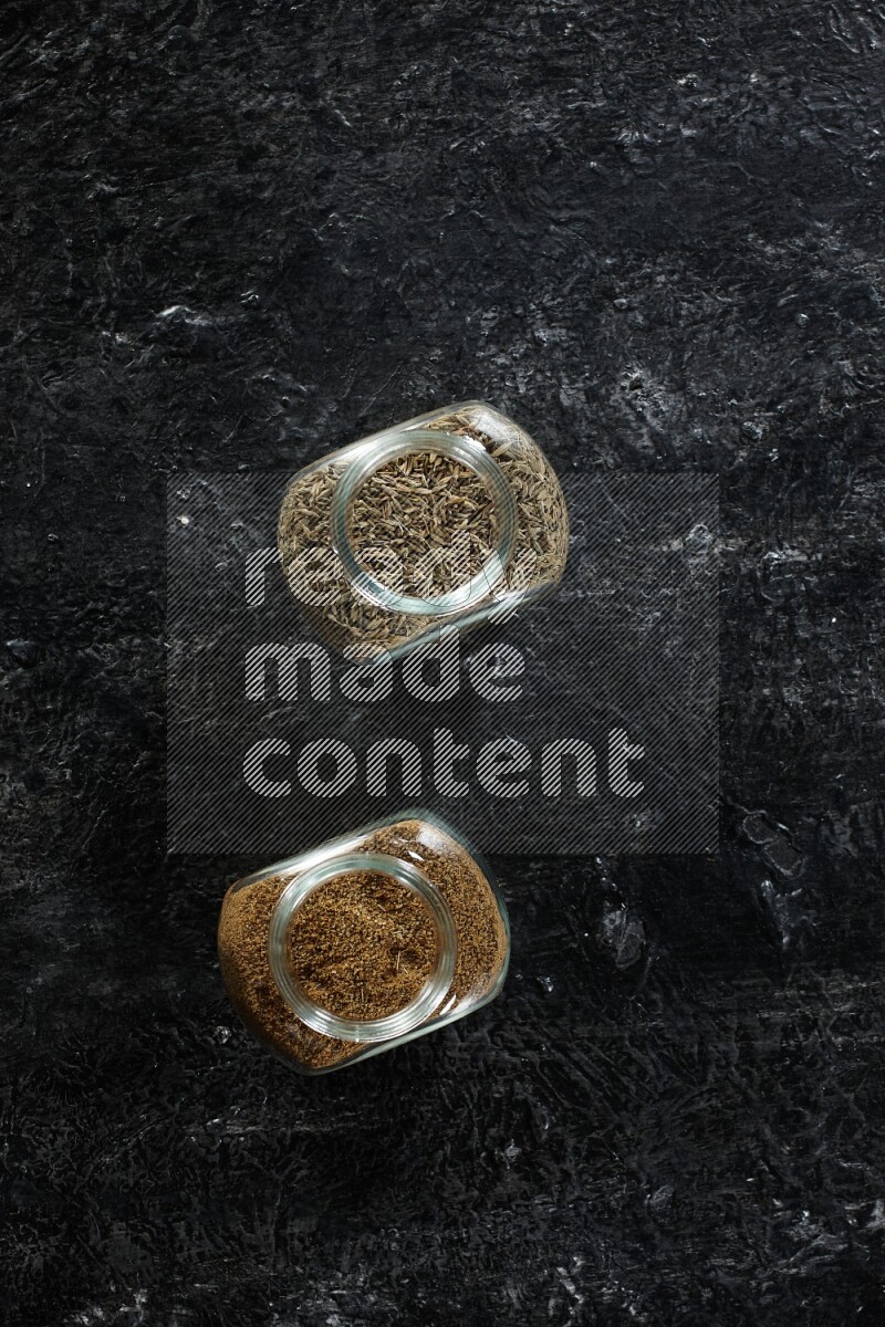 2 glass spice jars full of cumin powder and seeds on a textured black flooring