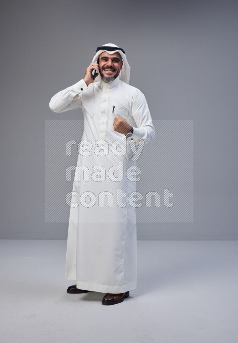 Saudi man Wearing Thob and white Shomag standing talking on phone on Gray background