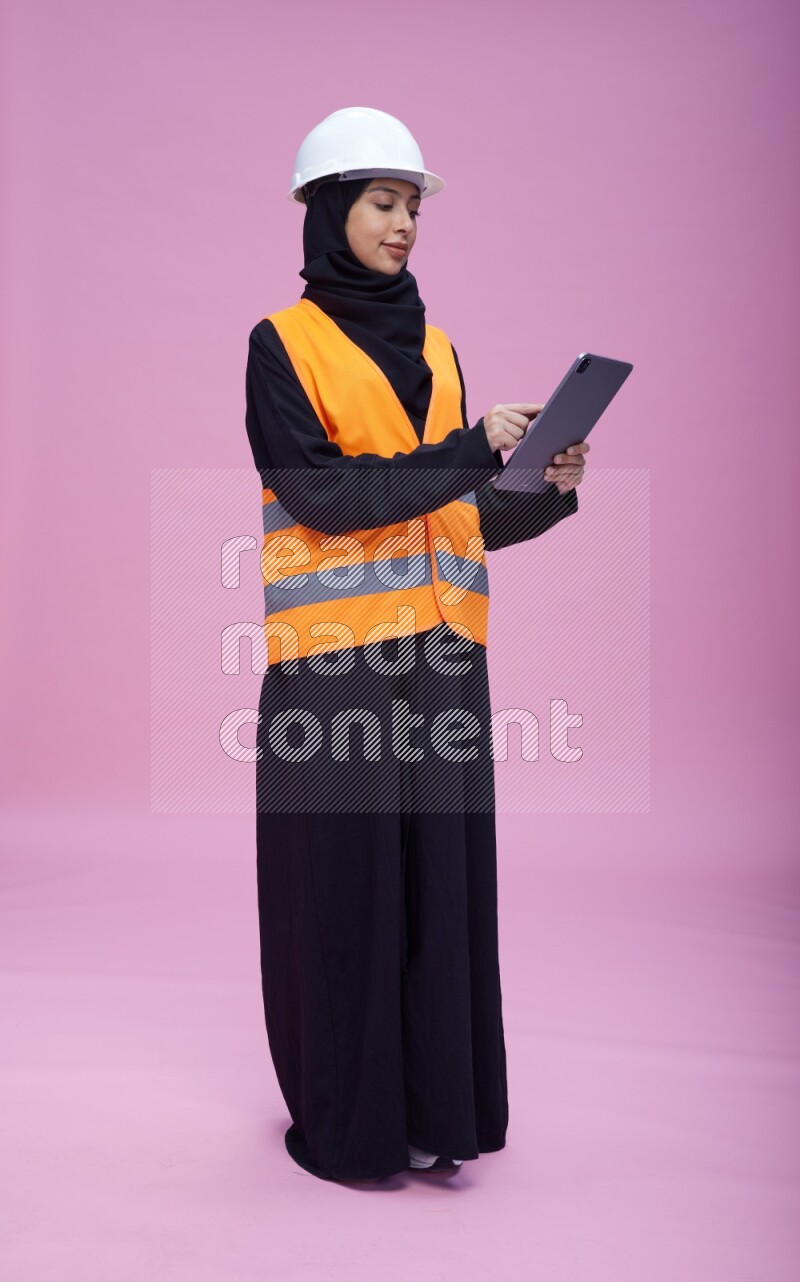 Saudi woman wearing Abaya with engineer vest and helmet standing working on tablet on pink background