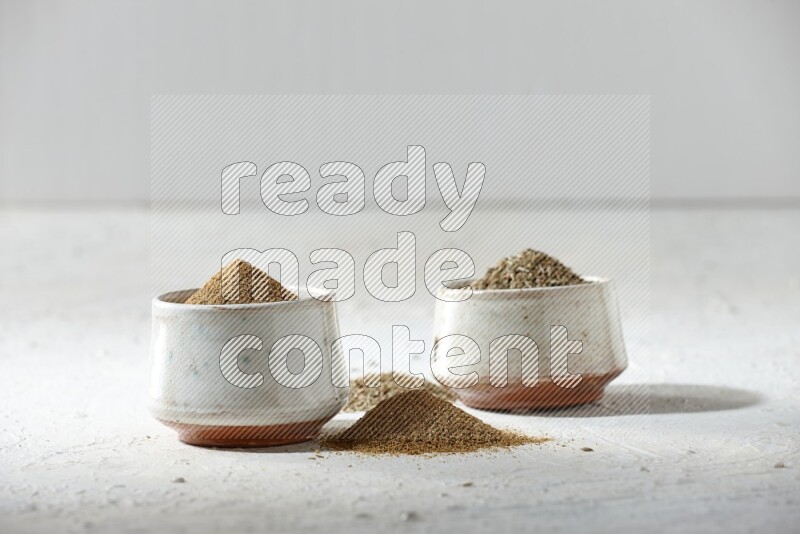 2 beige bowls full of cumin seeds and powder with spilled powder and seeds on textured white flooring