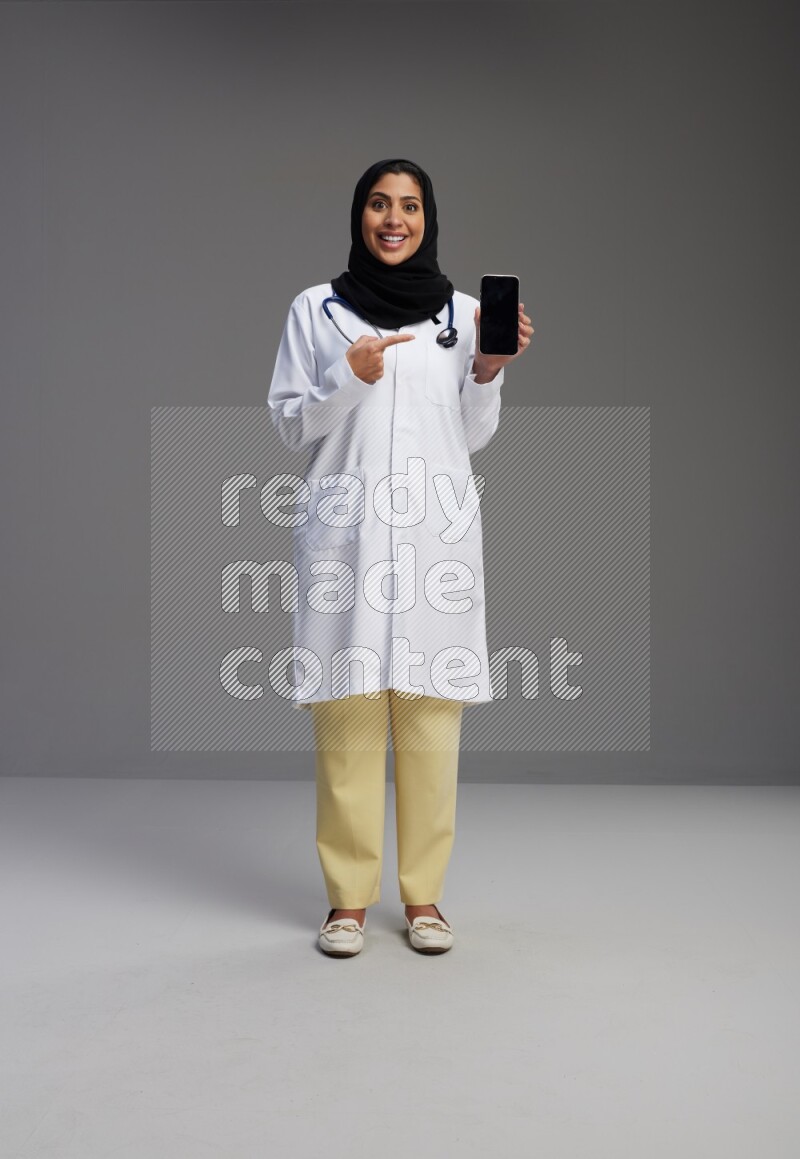 Saudi woman wearing lab coat with stethoscope standing showing phone to camera with sign in the back on Gray background