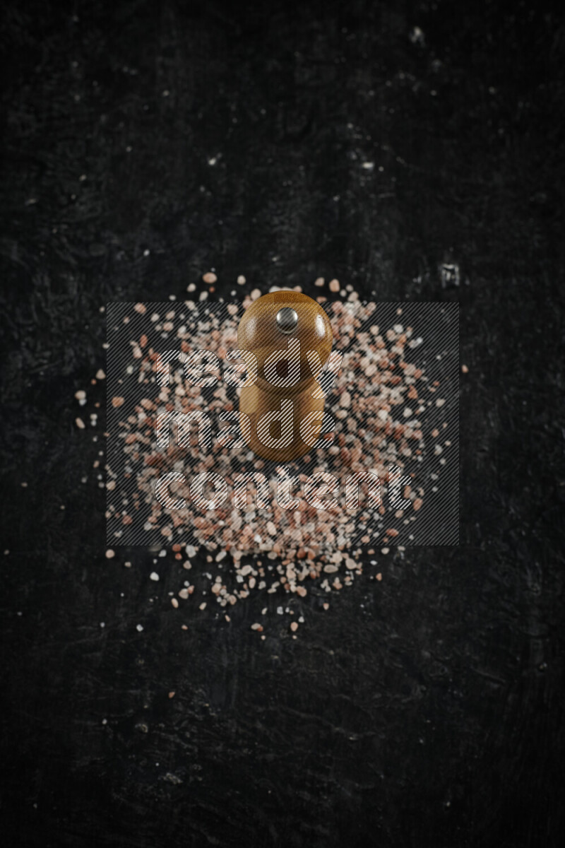 A wooden grinder standing upright and surrounded by coarse pink himalayan salt on black background