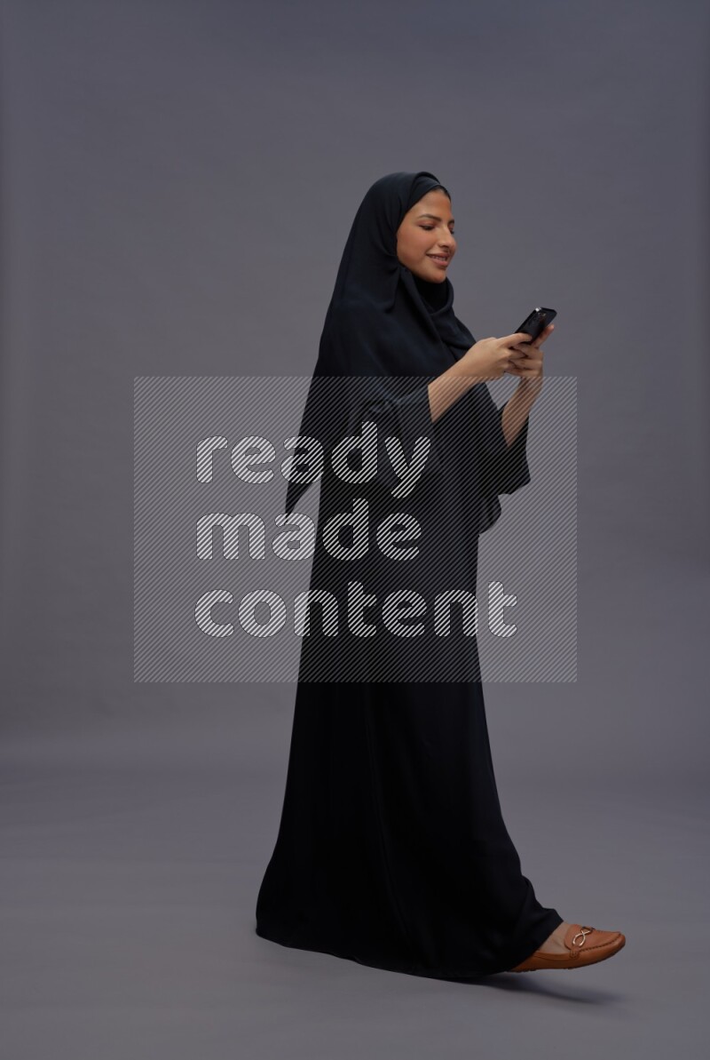 Saudi woman wearing Abaya standing texting on phone on gray background