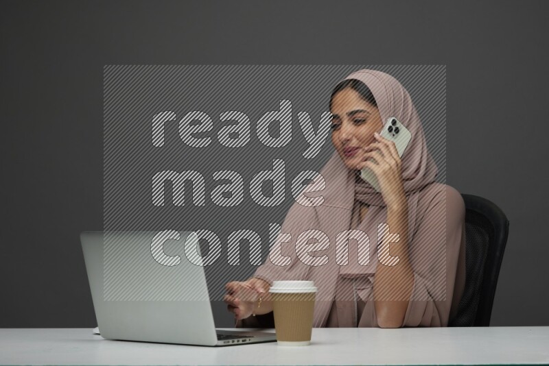 A Saudi woman Setting on her desk
 calling  on a Gray Background wearing Brown Abaya with Hijab