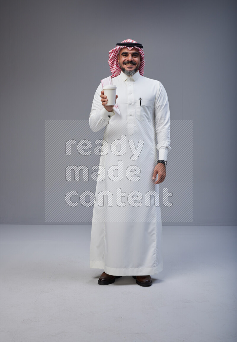 Saudi man Wearing Thob and red Shomag standing holding paper cup on Gray background