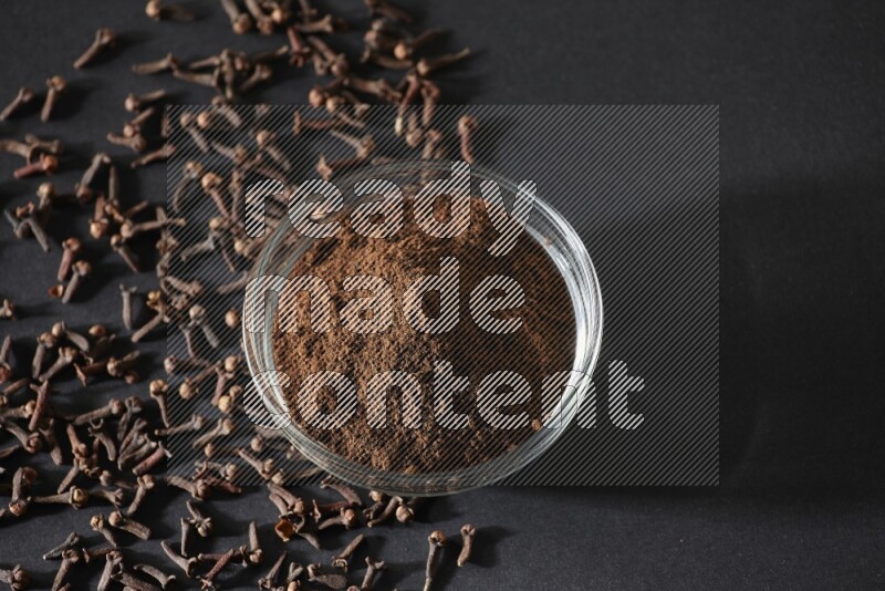 A glass bowl full of cloves powder with cloves grains spread on black flooring