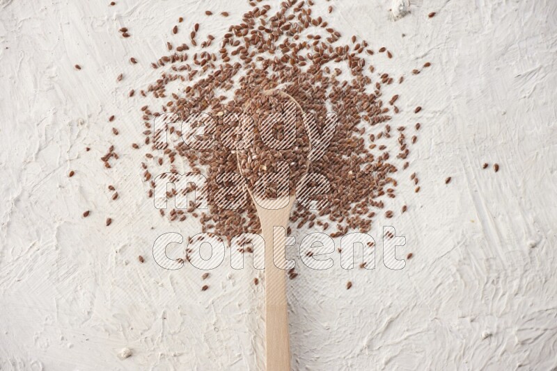 A wooden spoon full of flax seeds surrounded by flax seeds on a textured white flooring