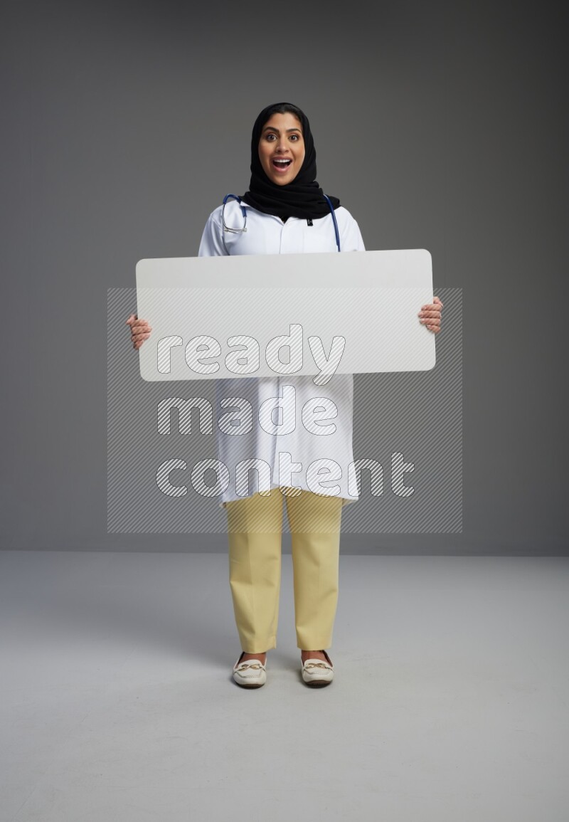 Saudi woman wearing lab coat with stethoscope standing holding board on Gray background