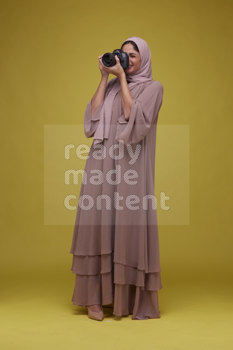 A woman Holding a Camera on a Yellow Background wearing Brown Abaya with Hijab