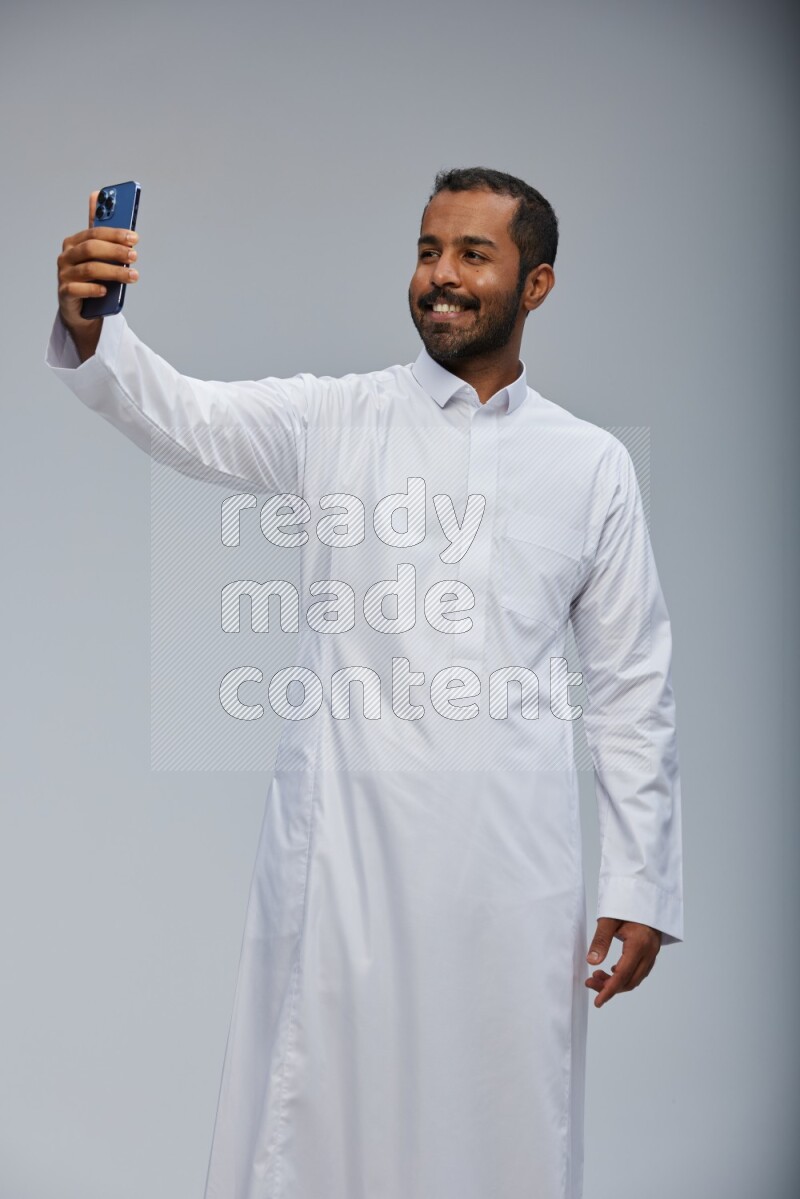 Saudi man wearing Thob standing taking selfie on Gray background