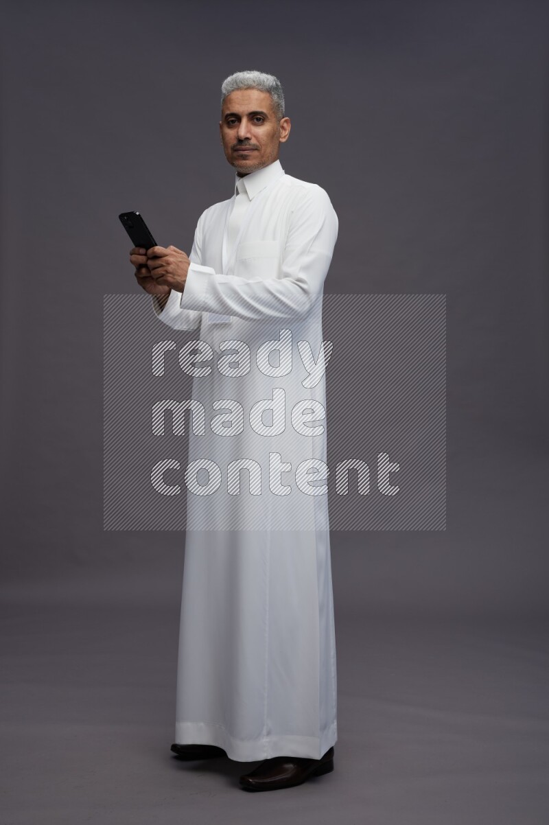Saudi man wearing thob with neck strap employee badge standing texting on phone on gray background