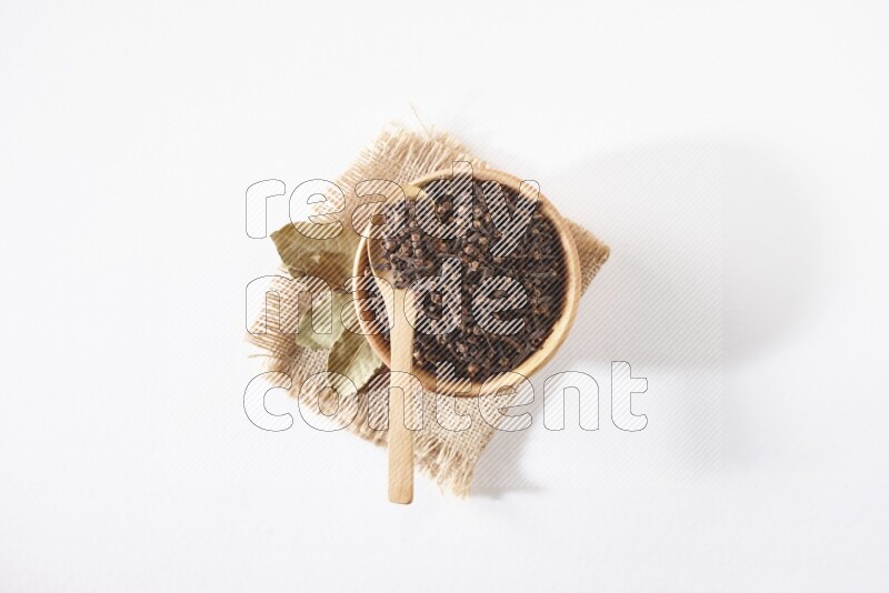 A wooden bowl and a wooden spoon full of cloves on a piece of burlap on a white flooring