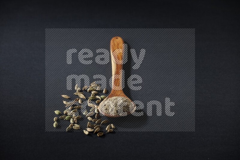 A wooden ladle full of cardamom powder and cardamom seeds beside it on black flooring