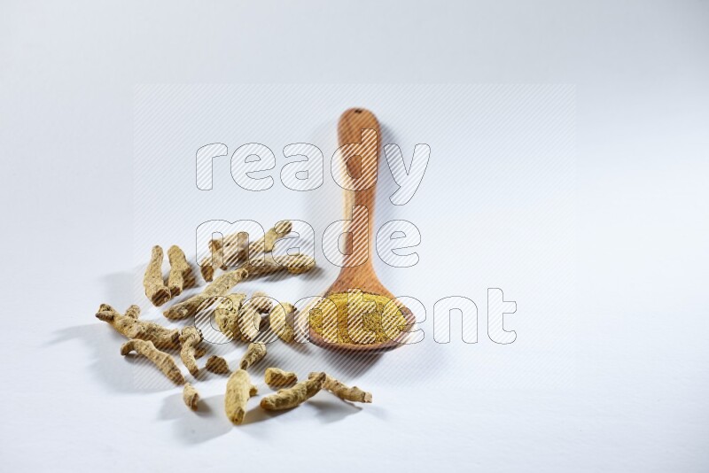A wooden ladle full of turmeric powder and dried turmeric fingers beside it on white flooring