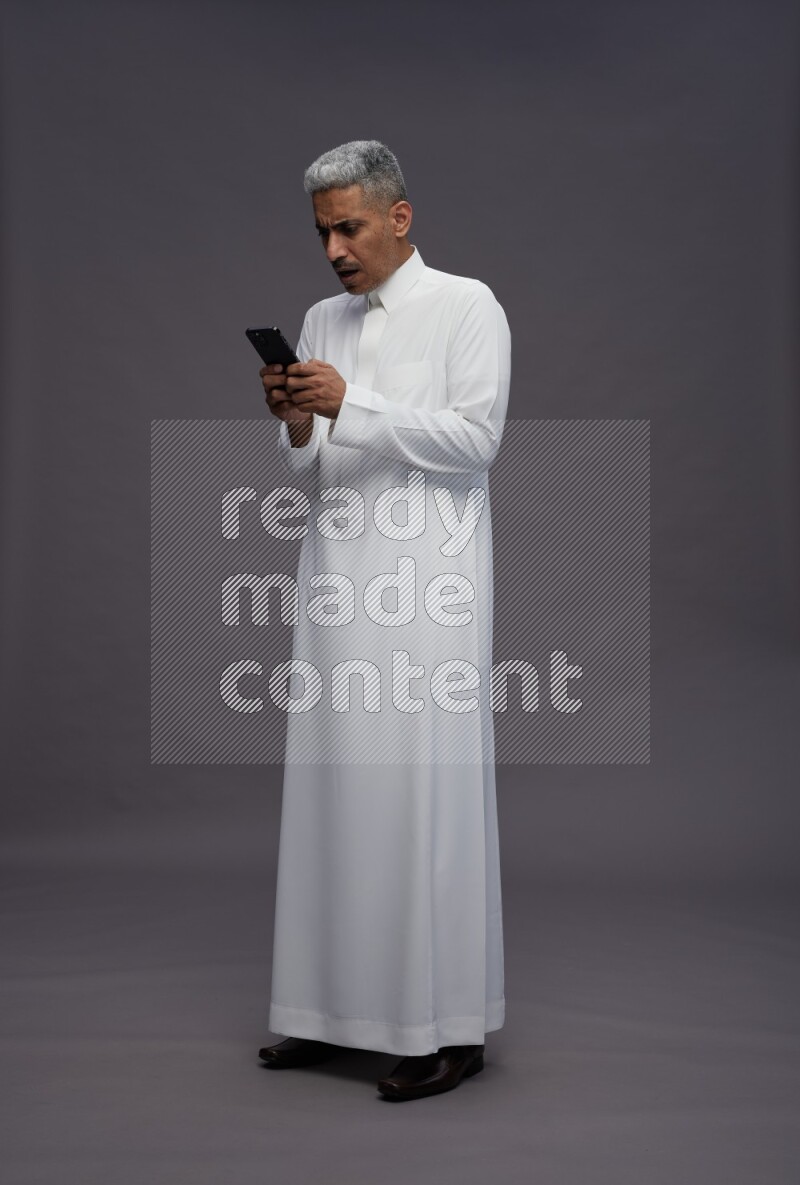 Saudi man wearing thob standing texting on phone on gray background