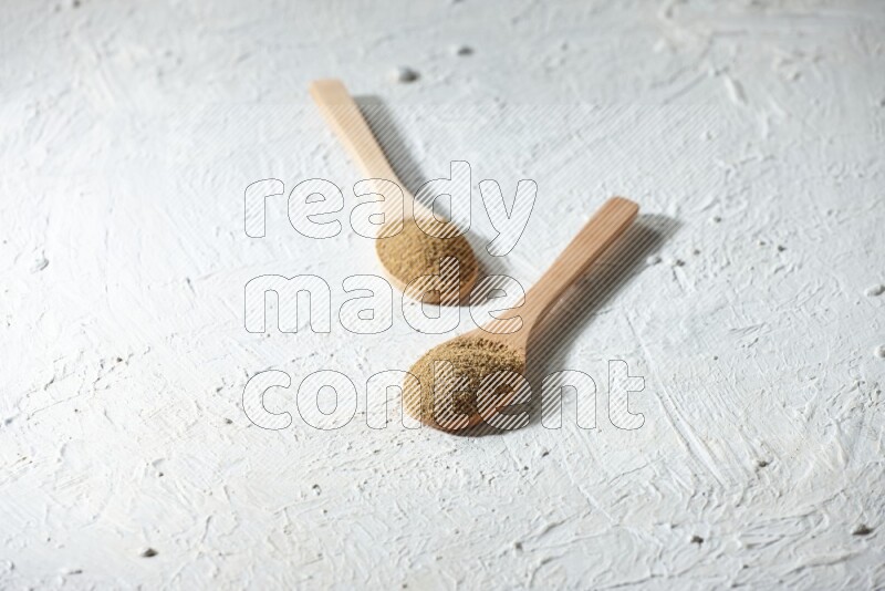 2 Wooden spoons full of cumin powder on a textured white flooring
