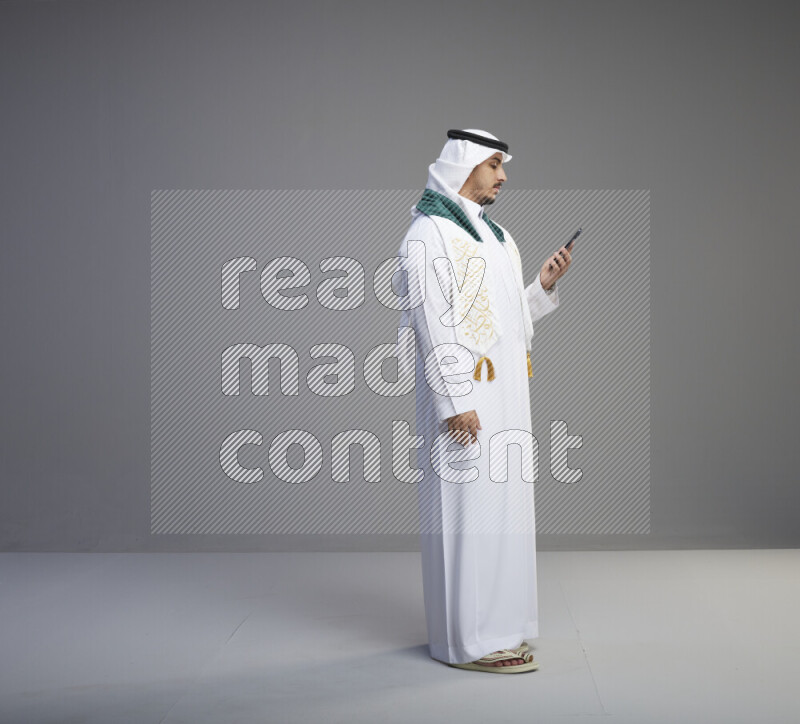 A saudi man standing wearing thob and white shomag with flag scarf on his neck talking on phone on gray background
