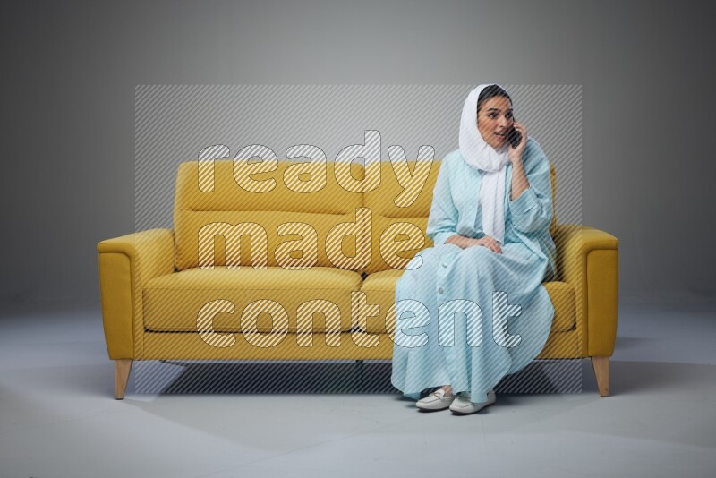 A Saudi woman wearing a light blue Abaya and a white head scarf sitting on a yellow sofa and talking in the phone while making different poses eye level on a grey background
