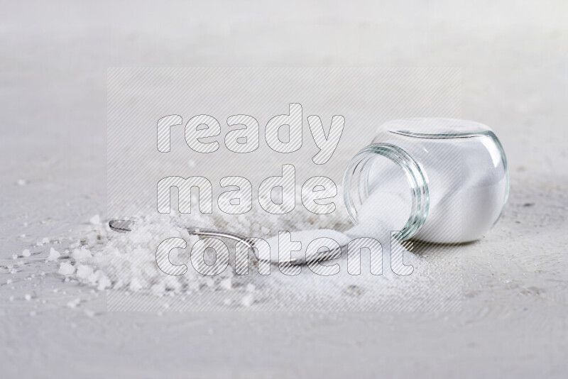 A glass jar full of table salt with some sea salt crystals beside it on a white background