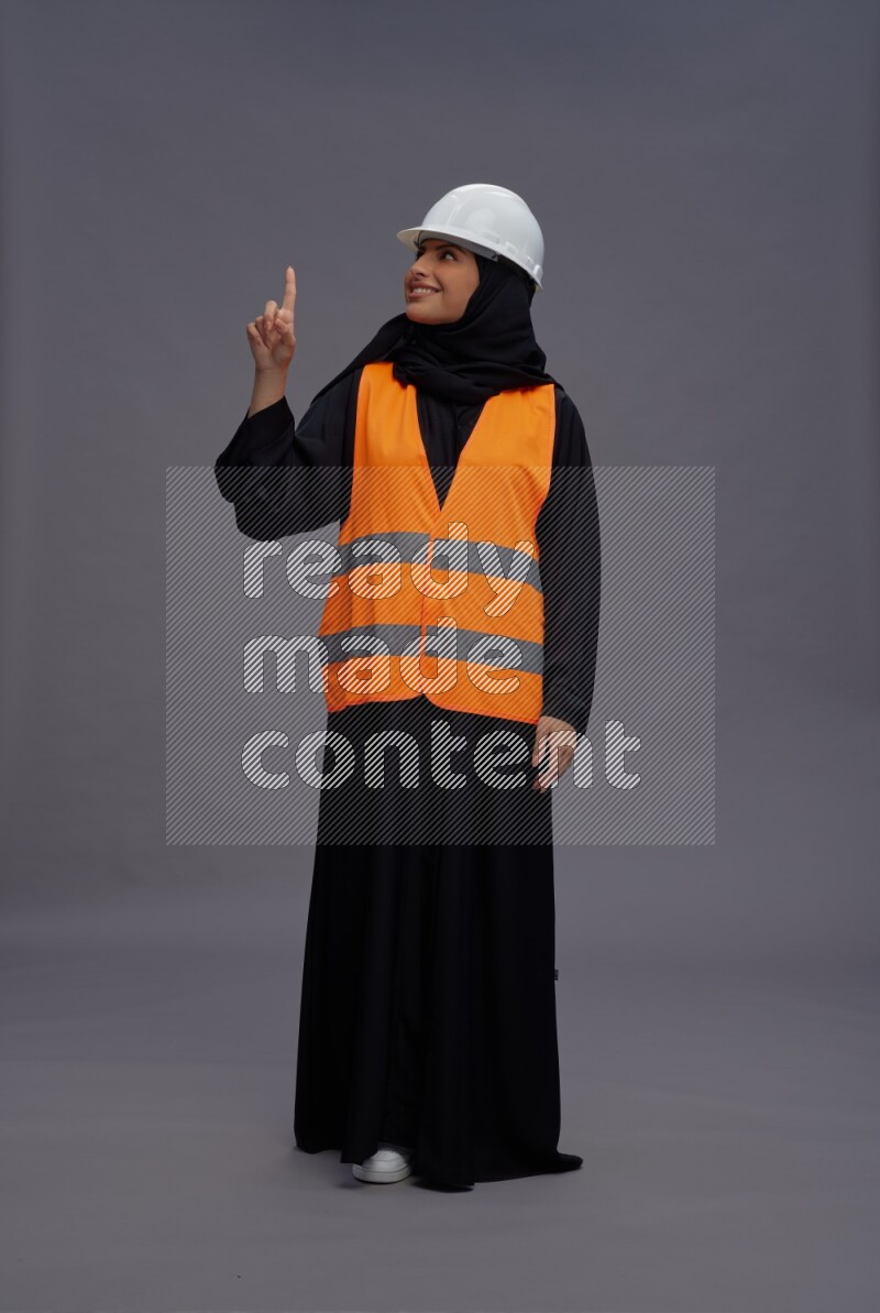 Saudi woman wearing Abaya with engineer vest standing interacting with the camera on gray background