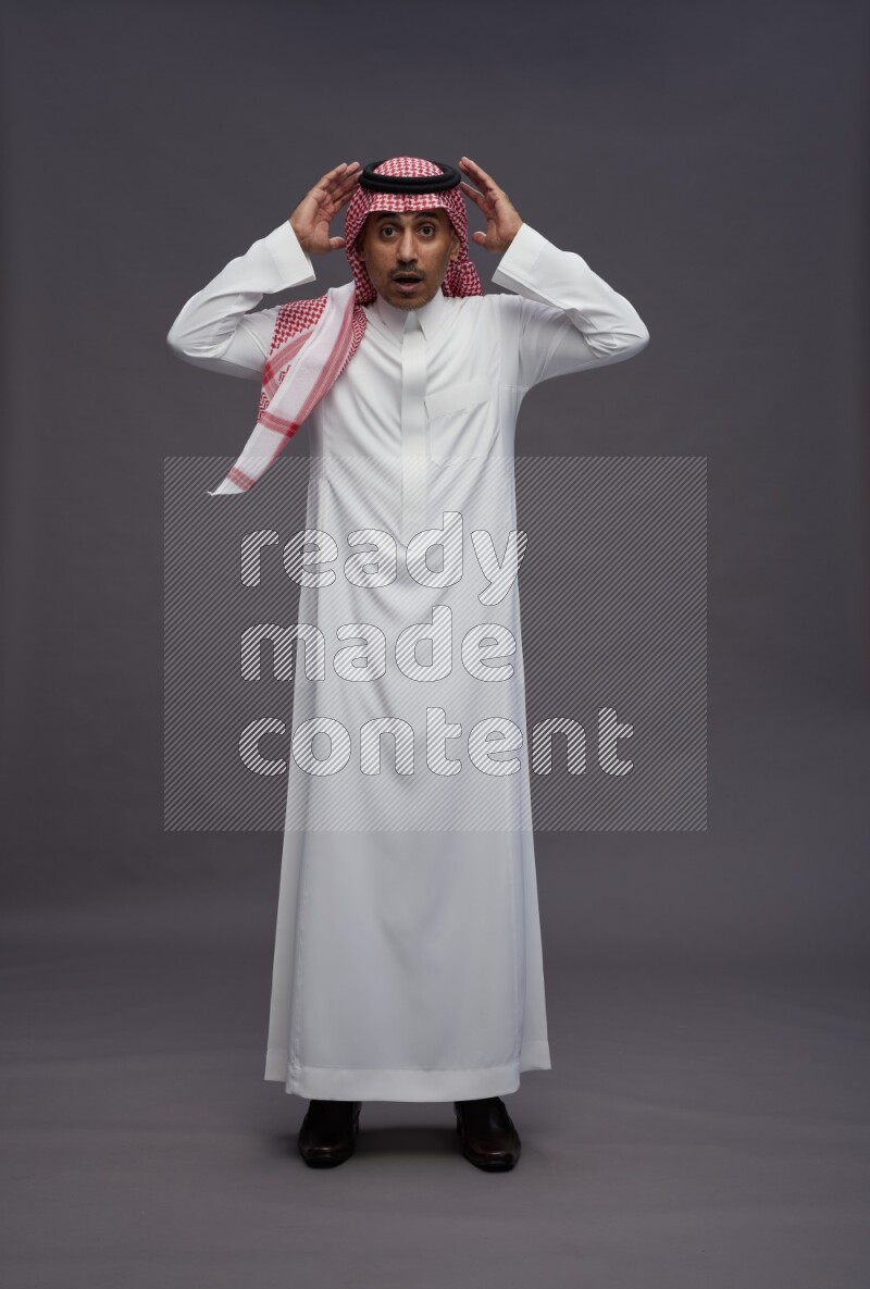 Saudi man wearing thob and shomag standing hands behind head on gray background