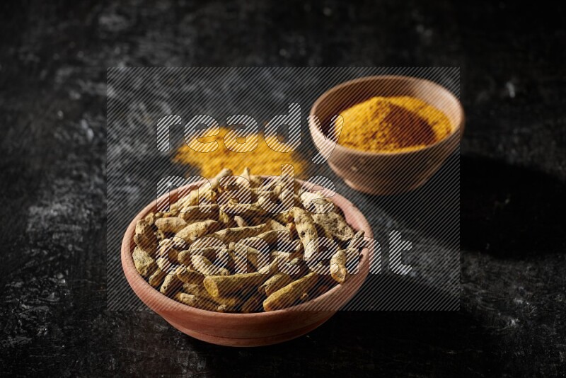 2 wooden bowls, one full of turmeric powder and the other full of dried turmeric whole fingers on a textured black flooring