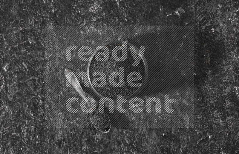 A black pottery bowl full of black seeds with a metal spoon full of the seeds on a textured black flooring