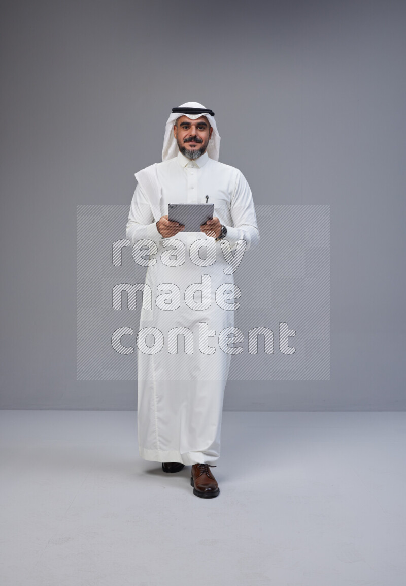 Saudi man Wearing Thob and white Shomag standing working on tablet on Gray background