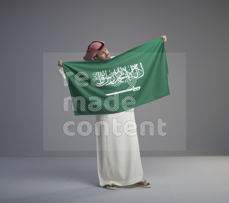 A saudi man standing wearing thob and red shomag holding big saudi flag on gray background