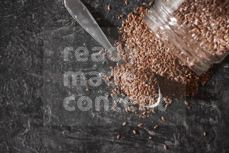 A glass jar full of flaxseeds flipped and seeds spread out with a metal spoon full of the seeds on a textured black flooring